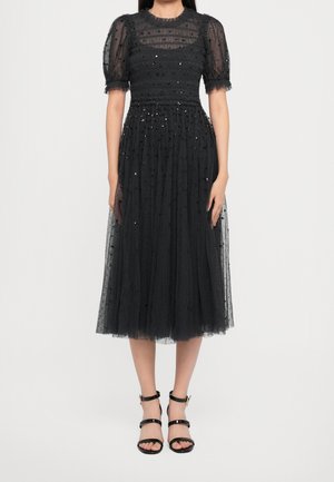 Woman wearing a black midi dress with puffed sheer sleeves, dotted pattern, and black ankle-strap heels, standing against a plain background.