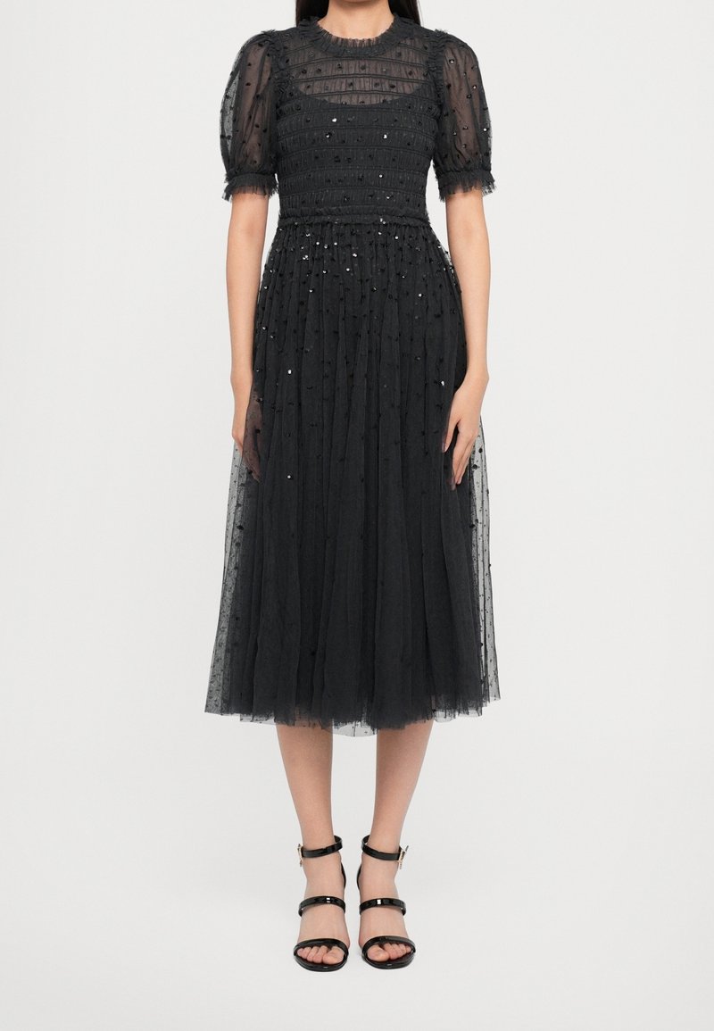 Femme portant une robe midi noire avec des manches bouffantes en tulle transparent, un motif à pois, et des escarpins noirs à bride autour de la cheville, debout devant un fond uni.