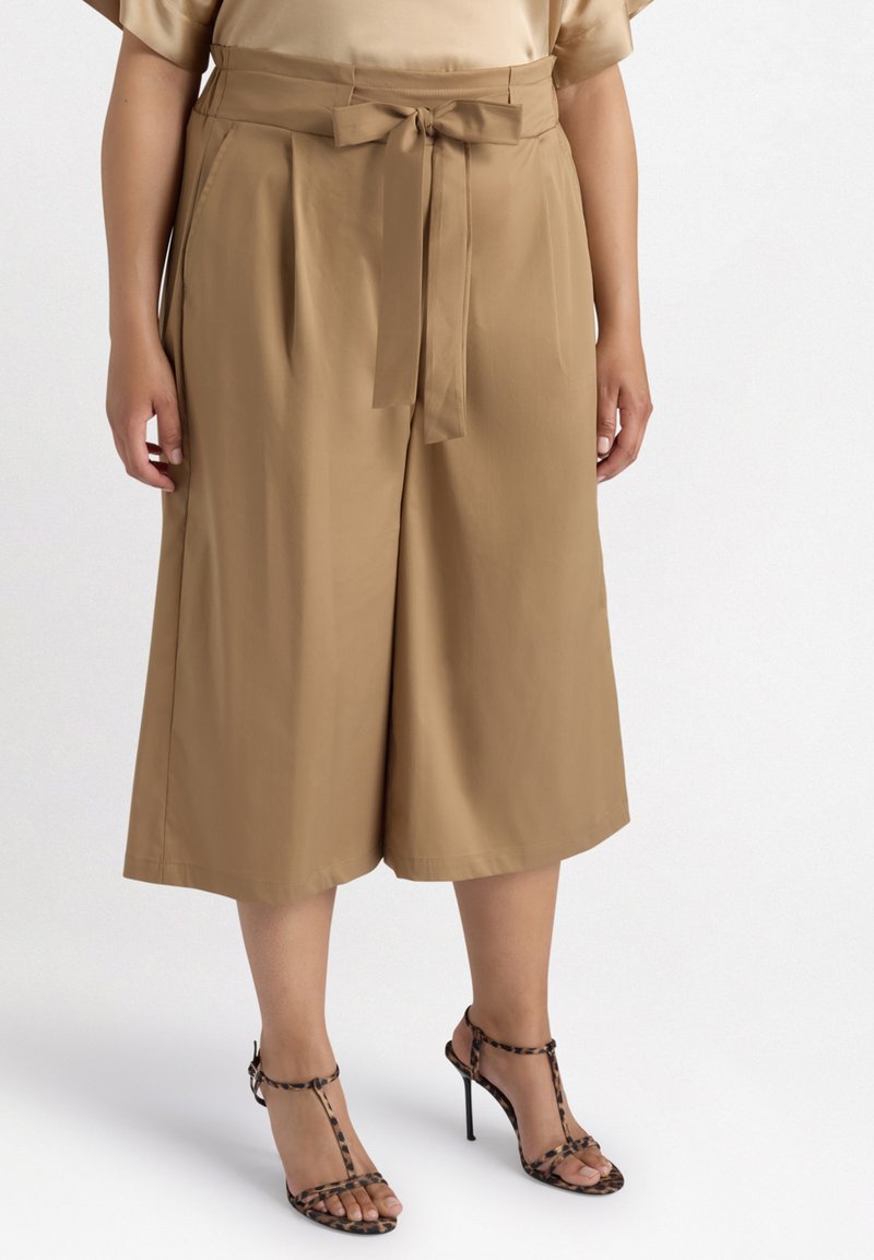 Culottes beiges en tissu lisse, dotés d'une taille haute avec une ceinture à nouer et des poches latérales, associés à des talons aiguilles noirs.