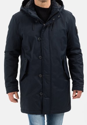 Homme portant un manteau d'hiver bleu marine à capuche avec des boutons, des poches zippées et un pull noir en dessous.