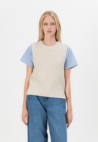 Pull sans manches côtelé crème avec un col rond, superposé à une chemise à manches courtes bleu et blanche à rayures ; porté avec un jean bleu.