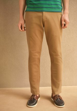 Kávové bavlnené nohavice so slim fit strihom a jemným elastanom. V páre s tmavými topánkami s hnedými akcentmi, so šnúrkami a bielymi podrážkami.
