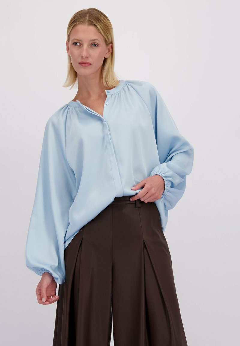 Lichtblauwe blouse met lange mouwen, een knoopsluiting aan de voorkant, geplooide schoudernaad en elastische manchetten, gecombineerd met bruine hoge taillebroek.
