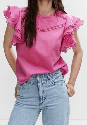 Frau trägt eine leuchtend pinke Bluse mit Rüschen und Spitzenbesatz, kombiniert mit hellblauen, hoch taillierten Jeans, die Hände in den Taschen vergraben.