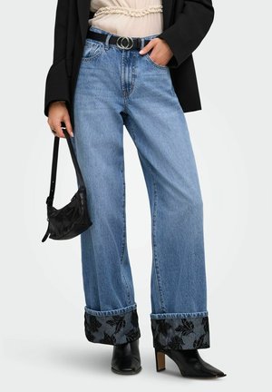 Femme portant un jean bleu à jambes larges avec des revers noirs à motif floral, des bottines noires à talons, un blazer noir, un haut crème et tenant un petit sac à main noir.