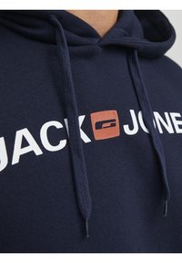 Sudadera azul oscuro con letras blancas "JACK" y "JONES." Presenta un logo cuadrado naranja y cordones, hecha de tela texturizada.