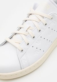 Zapatillas de piel blanca con cordones crema, detalles perforados y patrones sutiles del logo en el costado. Suela de goma texturizada.
