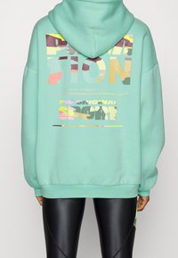 Felpa con cappuccio verde menta con stampe grafiche in testo multicolore sulla schiena. Presenta una vestibilità comoda e polsini a coste.