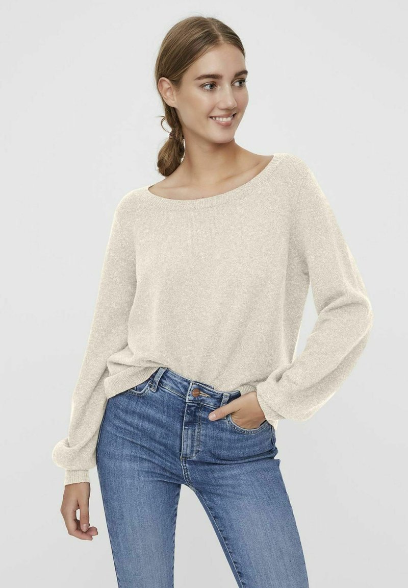 Pull en tricot couleur crème avec un large col, des manches longues et des poignets côtelés, associé à un jean taille haute bleu. Texture lisse.