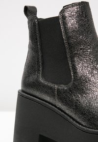 Botín negro con un upper de cuero metálico texturizado, paneles laterales elásticos y una gruesa suela de plataforma negra.