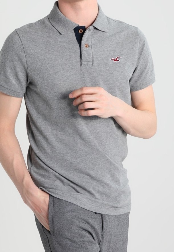 Homme portant un polo gris à manches courtes avec des boutons marron et un pantalon gris, main partiellement dans la poche et l'autre main en plein geste.