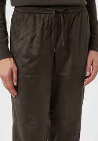 Close-up of brown soft fabric pants with elastic waistband and drawstring worn with matching long-sleeve top, hands relaxed at sides.