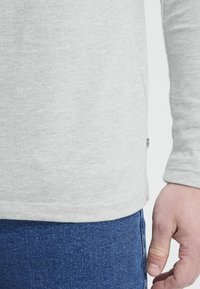 Light grey long-sleeve shirt with a soft texture, slightly rounded hem, and a small brand tag visible on the side, paired with blue denim.