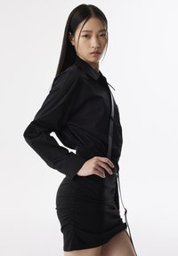 Camicia nera con bottoni, maniche lunghe e vita arricciata, abbinata a una gonna nera aderente con arricciature laterali e dettaglio con laccetto.