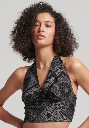 Mujer con cabello rizado vestida con un top halter negro con patrones de paisley blancos, de pie contra un fondo liso claro.