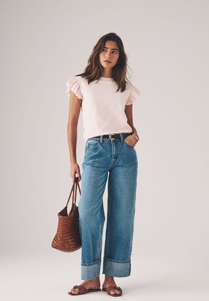 Vrouw staat met een lichtroze top met ruches aan de mouwen, wijde blauwgeribde jeans met brede boorden, bruine sandalen en houdt een gevlochten bruine handtas vast.