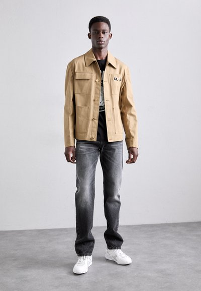 Light tan jacket with four front pockets, button closure, and a point collar. Worn over a black graphic t-shirt and paired with faded gray jeans.
