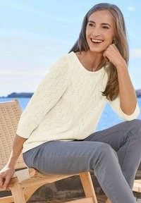 Hellcreme gestrickter Pullover mit weitem Halsausschnitt und Dreiviertelärmeln, kombiniert mit grauen Slim-Fit-Hosen, vor einer Küstenkulisse.