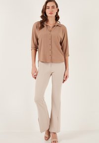 Brown button-up shirt with a collar, rolled sleeves. Paired with beige flared trousers featuring split hems and coordinating beige sandals.