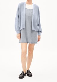 Vaaleansininen ribbineulottu neuletakki harmaan, taskullisen, hihattoman mekon pä ällä, jossa on repsottava helma. Asun täydentävät mustat slip-on-kengät.