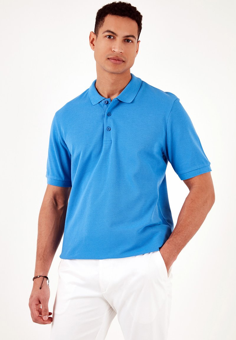 Camisa polo azul hecha de tejido texturizado, que cuenta con un collar, tres botones y mangas cortas. Combinada con pantalones blancos.
