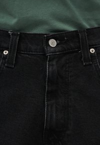 Κοντινό πλάνο στη μέση μαύρου τζιν με ασημένιο κουμπί και θηλιές για ζώνη, σε συνδυασμό με πράσινο υφασμάτινο τοπ.