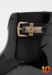 Botín negro de charol con panel elástico texturizado, banda metálica dorada y plateada con hebilla decorativa y detalle de logo.