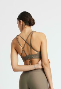 Mujer con cabello oscuro recogido en un moño, vistiendo un sujetador deportivo caqui con tirantes entrecruzados y leggings de talle alto a juego, mostrando un tatuaje en la parte baja de la espalda.