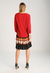 Blouse rouge à manches longues, associée à une robe à motifs avec un design en chevrons aux couleurs orange, crème et noir, et des talons noirs.