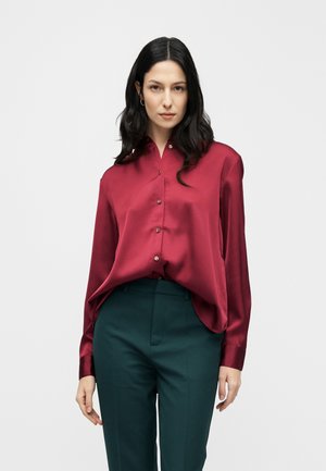 Pitkähiuksinen nainen, jolla on burgundinsävyinen silkkinen nappipaita ja tummanvihreät korkeavyötäröiset housut, yksinkertaisen taustan edessä.