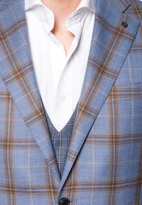 Jasnoniebieska marynarka w kratę z brązowymi akcentami, z teksturowanej tkaniny, z czarnym guzikiem i białą koszulą pod spodem.