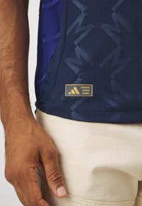 Pánský navy sportovní dres, texturovaná látka s jemným vzorem cik-cak, s zlatou štítkovou etiketou s textem "AUTENTICKÝ LICENCOVANÝ PRODUKT".