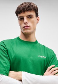 T-shirt de algodão verde com decote redondo e mangas curtas. Apresenta um pequeno logotipo branco na área do peito. Textura suave.