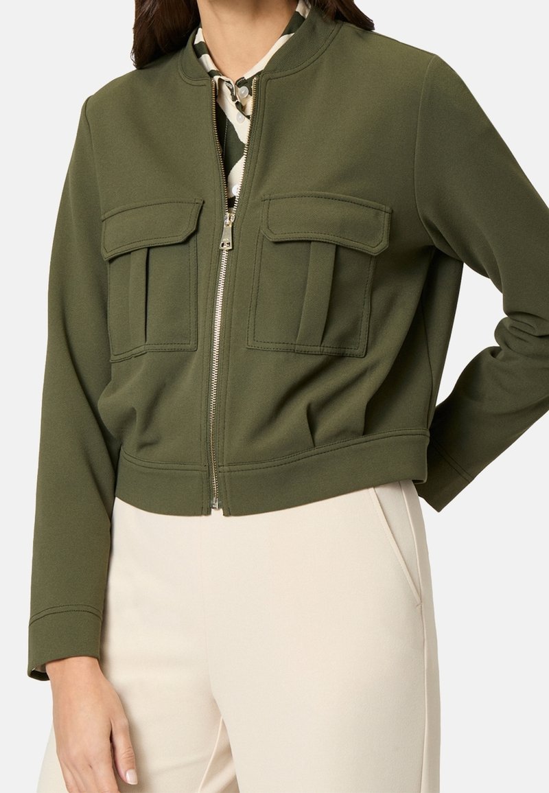 Gekürzte olivgrüne Jacke aus glattem Stoff, mit durchgehendem Reißverschluss vorne, zwei Brusttaschen mit Patten und gerippten Bündchen.