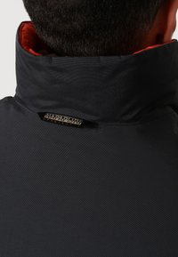 Homme portant une veste noire avec un col montant doublé de rouge et un logo Napapijri à l'arrière du col.