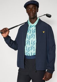Veste marine sur une chemise polo à motifs bleu clair avec design LS1, pantalon noir et casquette marine. Un club de golf repose sur son épaule.