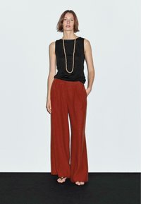 Black sleeveless top and wide-legged rust pants made of textured fabric; accessorized with a long beaded necklace, against a plain background.
