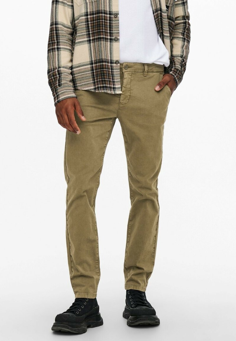 Olivengrønne bomuldsbukser med lige snit, båret med en beige og mørkternet skjorte. Sorte snørebåndsstøvler fuldender outfittet.