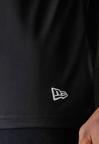 Schwarzes Sportshirt aus strukturiertem Stoff. Gesticktes weißes Logo in der linken unteren Ecke. Einfaches Design mit einer figurbetonten Silhouette.