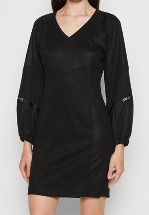 Robe mini noire à manches longues avec col en V, en tissu légèrement brillant et manches bouffantes comportant de petits détails de texte blanc près des coudes.