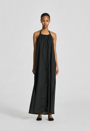 Femme debout, face à nous, portant une robe maxi noire sans manches à boutons avec un col halter et des chaussures plates noires sur un fond uni.