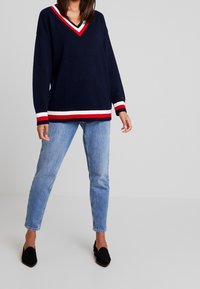 Marineblauer Strickpullover mit V-Ausschnitt, ausgestattet mit roten und weißen Streifen am Saum und an den Bündchen. Kombiniert mit hellblauen schmal geschnittenen Jeans und schwarzen Loafers.