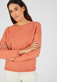 Pull en tricot couleur corail avec un motif texturé sur la poitrine, col rond, et manches longues avec des poignets côtelés.