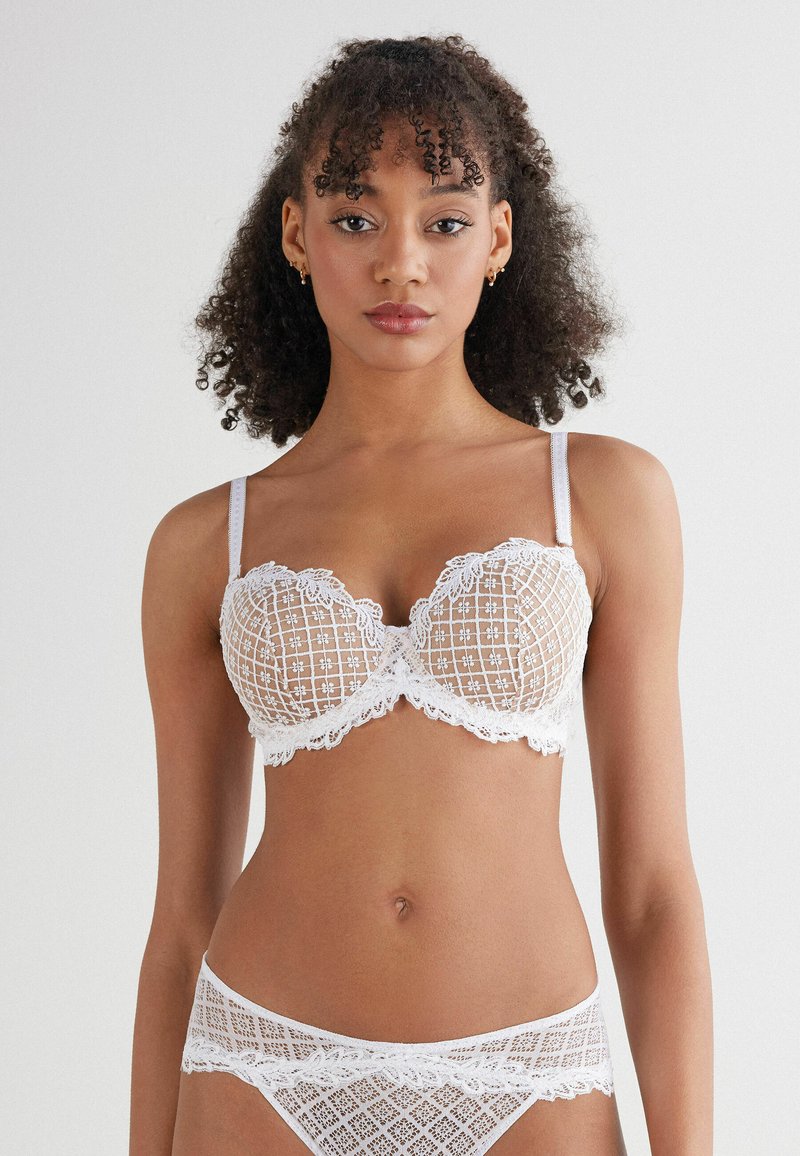 Vit spets-bh med rutmönster och blommor. Har byglar, justerbara axelband och en matchande genomskinlig spetslinning.