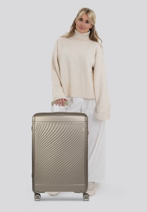 Silver hard-shell suitcase with wavy texture, two wheels, and an extendable handle. Displayed next to a person in light-colored clothing.