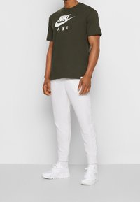 Olivově zelené tričko Nike s bílým logem, doplněné o bílé tepláky s bočními pruhy a bílé sportovní boty. Jednoduchý, neformální vzhled.