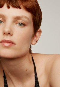 Pendiente de aro plateado con un acento azul, colocado en la oreja izquierda. El sujeto tiene el cabello corto de color castaño rojizo y un tono de piel neutral, con maquillaje mínimo.