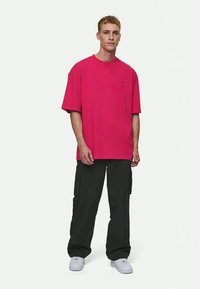 T-shirt rosa choque de manga curta com um corte relaxado, combinado com calças cargo pretas. O modelo usa ténis brancos, de pé contra um fundo claro.