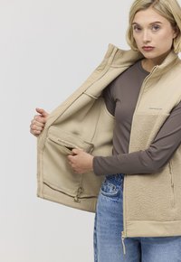 Beige Fleece-Weste mit Reißverschluss vorne, strukturiertem Körper und glatten Schultern; verfügt über eine Innentasche mit dem Etikett "MADE FOR EXPERIENCES".