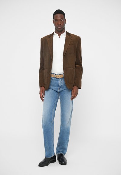 Brun fløjlsblazer over en hvid skjorte, parret med lyseblå jeans og sorte sko. Blazeren har synlige lommer og tekstiler med struktur.